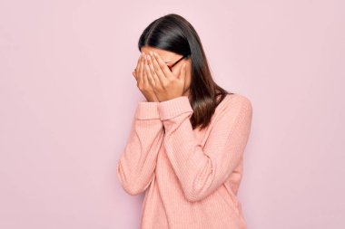 Genç, güzel, esmer bir kadın pembe arka planda günlük süveter ve gözlük takıyor. Ağlarken yüzünü ellerle kapatan üzgün bir ifade var. Depresyon kavramı.