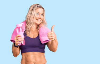 Orta yaşlı, sarışın, spor kıyafeti giyen, havluyla gezen, elinde su şişesi tutan mutlu ve pozitif gülümseyen, başparmağını kaldıran, mükemmel ve onay işareti yapan bir kadın. 