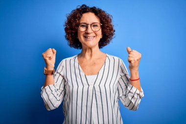 Orta yaşlı, güzel, kıvırcık saçlı, sıradan çizgili tişörtlü bir kadın izole edilmiş arka planda şaşırmış ve şaşırmış bir şekilde kolları açık, gözleri açık bir şekilde başarıyı kutluyor. Kazanan konsept.