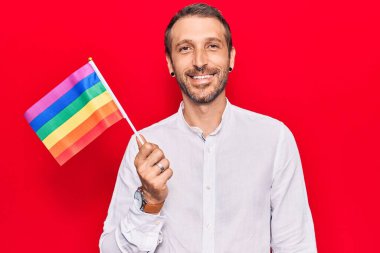 Elinde gökkuşağı lgbtq bayrağı tutan genç yakışıklı adam pozitif ve mutlu görünüyor ve kendine güvenen bir gülümsemeyle dişlerini gösteriyor. 