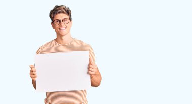 Boş pankart tutan yakışıklı genç adam pozitif ve mutlu görünüyor. Kendine güvenen bir gülümsemeyle ayakta duruyor ve dişlerini gösteriyor. 