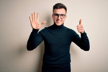 Gözlüklü, sıradan bir süveter giyen genç, yakışıklı beyaz bir adam izole edilmiş arka planda altı numarayı işaret ederken kendinden emin ve mutlu bir şekilde gülümsüyor..