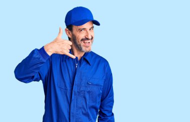 Middle age handsome man wearing mechanic uniform smiling doing phone gesture with hand and fingers like talking on the telephone. communicating concepts.  clipart
