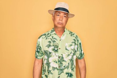 Plaj tatilinde yaz şapkası ve çiçekli gömlek giyen orta yaşlı, kır saçlı bir adam. Yüzünde ciddi bir ifadeyle rahatlamış. Basit ve doğal bir şekilde kameraya bakıyorsun..