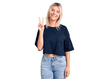 Gündelik tişört giyen genç ve güzel sarışın kadın iki numaralı parmaklarıyla işaret ederken kendine güvenen ve mutlu bir şekilde gülümsüyor.. 