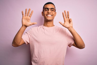 Yakışıklı Afro-Amerikalı adam pembe arka plan üzerinde günlük tişört ve gözlük takıyor ve dokuz numarayı gösterirken kendine güvenen ve mutlu bir şekilde gülümsüyor..