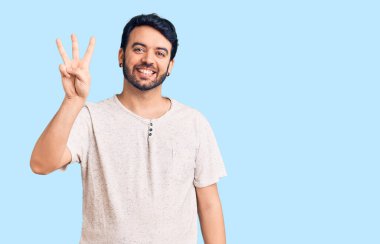 Gündelik kıyafetler giyen genç İspanyol adam üç numarayı gösterip işaret ederken kendinden emin ve mutlu gülümsüyor.. 