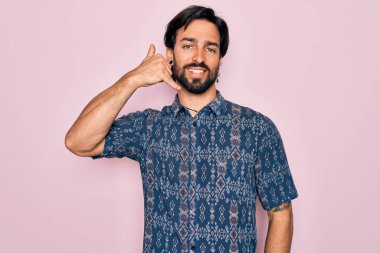 Pembe arka planda hippi tarzı giyinen yakışıklı İspanyol bohem adam telefonla el ve parmaklarıyla telefonla konuşur gibi gülümsüyor. İletişim kavramları.