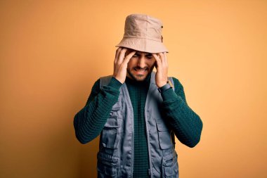 Tatilde sakallı yakışıklı bir turist sarı arka planda kaşif şapkası takıyor. Ağrı ve migren yüzünden çaresiz ve stresli. Ellerini başının üstüne koy..