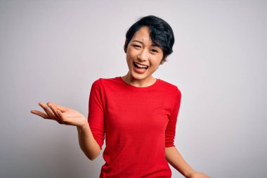 Genç, güzel Asyalı bir kız, günlük kırmızı tişört giyip izole edilmiş beyaz arka planda dikiliyor. Neşeli gülümsüyor, kollarını açıyor, dostça karşılıyor, olumlu ve kendinden emin selamlıyor.