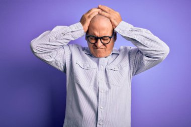 Orta yaşlı, beyaz, çizgili gömlek ve mor arka planda gözlük takan yakışıklı adam acı ve migren yüzünden çaresiz ve stresli baş ağrısından muzdarip. Ellerini başının üstüne koy..