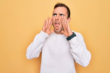 Mavi gözlü, sıradan bir süveter giyen, sarı arka planda duran genç, yakışıklı bir adam elleri ağzında sinirli bir şekilde bağırıyor.