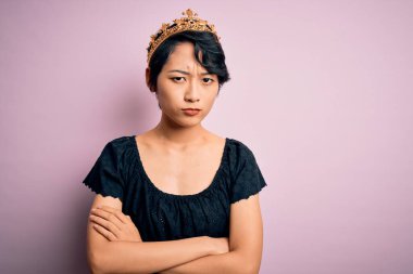 Soyutlanmış pembe arka plan üzerinde kralın altın tacını takan genç, güzel Çinli kadın şüpheci ve gergin, yüzünde çapraz kollu tasvip etmeyen ifade. Negatif kişi.