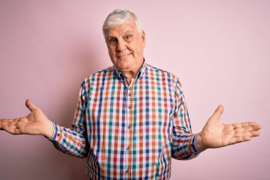 Üst düzey yakışıklı, parlak gömlekli, pembe arka planda izole edilmiş gülümseyen, her iki elini de açık gösteren, sunan ve reklam karşılaştırma ve denge