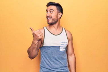 Sakallı, kolsuz tişörtlü, sarı arka planda duran genç yakışıklı adam baş parmağını yana kaldırıyor ve ağzı açık gülümsüyor.