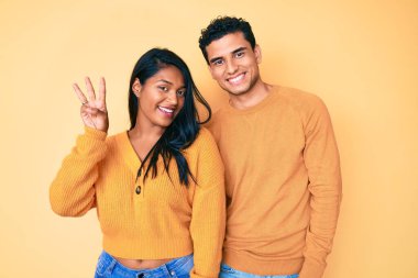 Güzel Latin genç çift, günlük kıyafetlerini giyip üç numaralı parmaklarını göstererek mutlu ve kendinden emin bir şekilde gülümsüyorlar.. 