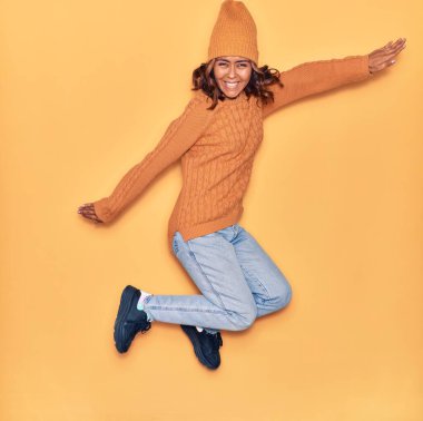 Yün şapka ve süveter giyen, mutlu gülümseyen genç, güzel Latin bir kadın. Yüzünde tebessümle zıplıyor ve kollarını izole edilmiş sarı arka planda açıyor.