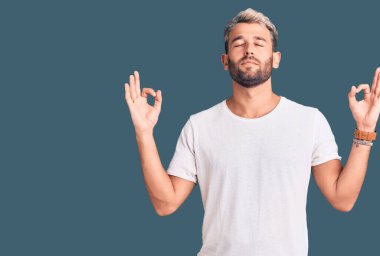 Gündelik tişört giyen, mutlu yüzlü gülümseyen ve baş parmağıyla yüzünü gösteren genç, yakışıklı sarışın bir adam.. 