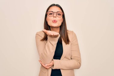 Young beautiful brunette businesswoman wearing jacket and glasses over white background looking at the camera blowing a kiss with hand on air being lovely and sexy. Love expression. clipart