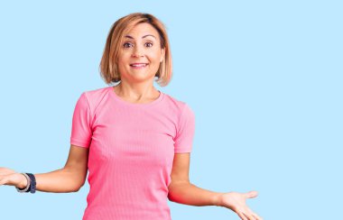 Spor kıyafetleri giyen genç sarışın kadın neşeli ve kollarını açmış gülümsüyor. Hoş geldin, olumlu ve kendinden emin selamlar. 