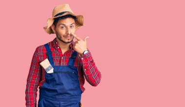 Yakışıklı Latin Amerikalı, giyimli, tamirci üniforması giyen, el parmağı ve burnuyla işaret eden, neşeli gülümseyen genç adam. güzellik konsepti 