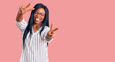 Gündelik kıyafetler ve gözlük takan genç Afro-Amerikan kadın iki elini de zafer işareti yaparken göstererek gülümsüyor. İki numara.. 