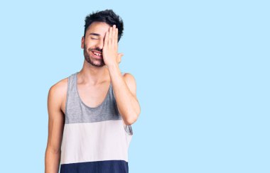 Latin asıllı genç adam gündelik giysiler giyiyor. Bir gözünü eliyle kapatıyor. Yüzünde kendine güvenen bir gülümseme ve sürpriz bir duygu var.. 