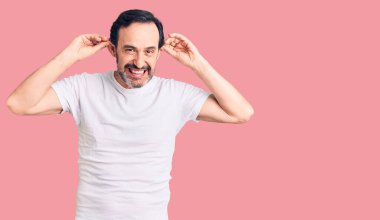 Orta yaşlı yakışıklı bir adam günlük tişört giyiyor gülümsüyor kulakları parmaklarıyla çekiyor, komik bir jest. deneme sorunu 