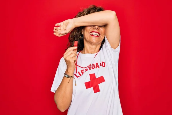 Middle age senior summer lifeguard woman holding whistle over red background Smiling cheerful playing peek a boo with hands showing face. Surprised and exited