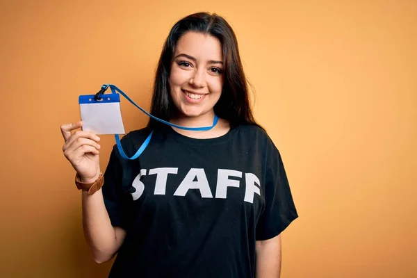 Young brunette worker woman wearing staff t-shirt as uniform showing id card with a happy face standing and smiling with a confident smile showing teeth