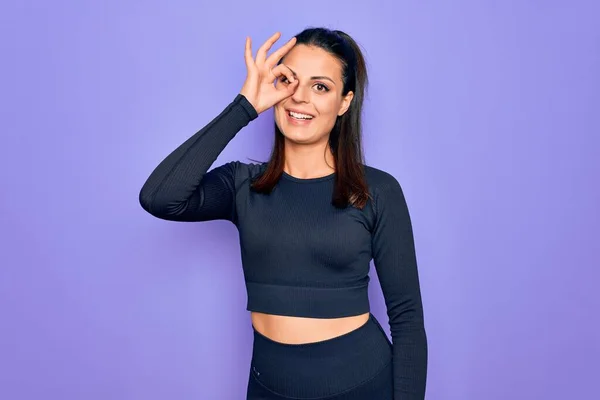 Young beautiful brunette sporty woman wearing casual sportswear over purple background doing ok gesture with hand smiling, eye looking through fingers with happy face.