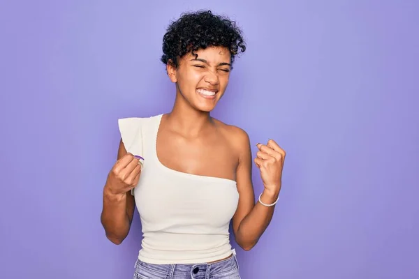 年轻美丽的非洲裔美国女人 穿着紫色背景的休闲T恤 非常快乐而兴奋地举起双臂 做着获胜的手势 微笑着 尖叫着要成功 庆祝概念 — 图库照片