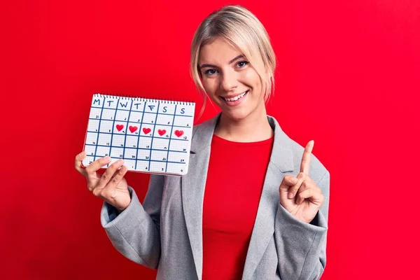 Piękna Blondynka Kontrolująca Miesiączkę Trzymając Kalendarz Czerwonym Tle Uśmiechając Się — Zdjęcie stockowe