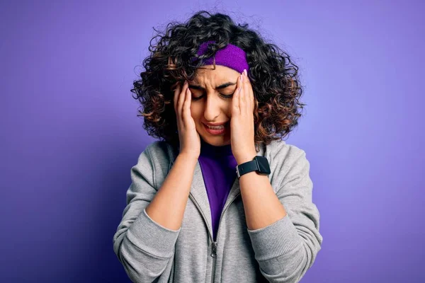 Beautiful curly arab sportswoman doing sport wearing sportswear over purple background with hand on head for pain in head because stress. Suffering migraine.