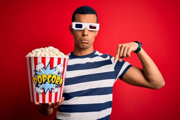 Young handsome african american man watching movie using 3d glasses eating popcorns Pointing down looking sad and upset, indicating direction with fingers, unhappy and depressed.