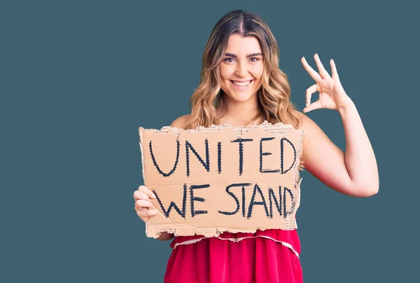 Young caucasian woman holding united we stand banner doing ok sign with fingers, smiling friendly gesturing excellent symbol