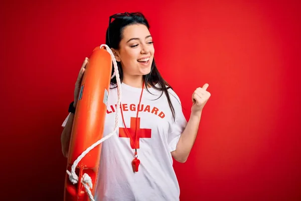 Young lifeguard woman wearing secury guard equipent holding rescue float over red background pointing and showing with thumb up to the side with happy face smiling