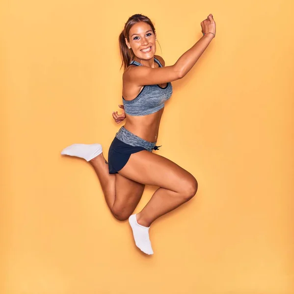 スポーティーな服を着て若い美しいヒスパニック系スポーツ女性は幸せな笑顔 孤立した黄色の背景の上に顔に笑顔でジャンプ — ストック写真