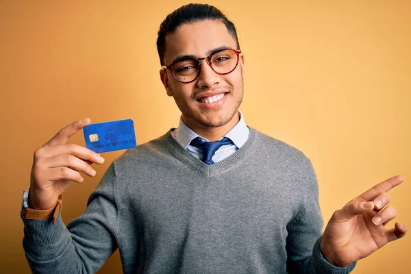 Young brazilian businessman holding credit card money over isolated yellow background very happy pointing with hand and finger to the side