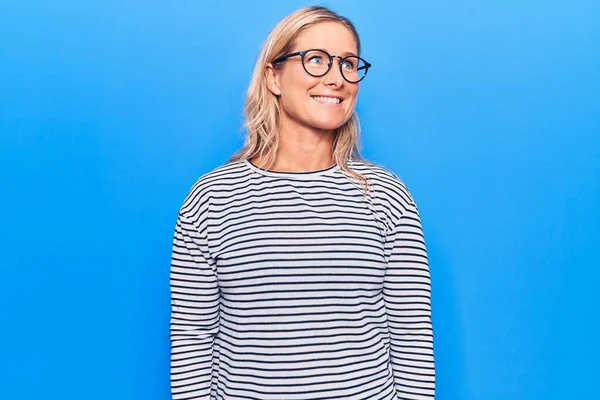 Orta yaşlı, beyaz, çizgili süveter ve gözlük takan, yüzünde gülümseme, doğal bir ifade olan sarışın kadın. Kendinden emin gülen. 