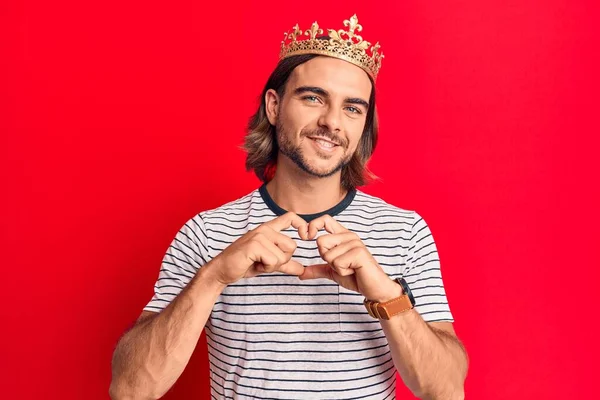 Young handsome man wearing prince crown smiling in love doing heart symbol shape with hands. romantic concept.