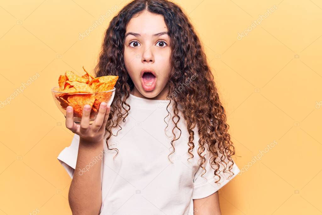 Beautiful kid girl with curly hair holding nachos potato chips scared and amazed with open mouth for surprise, disbelief face 