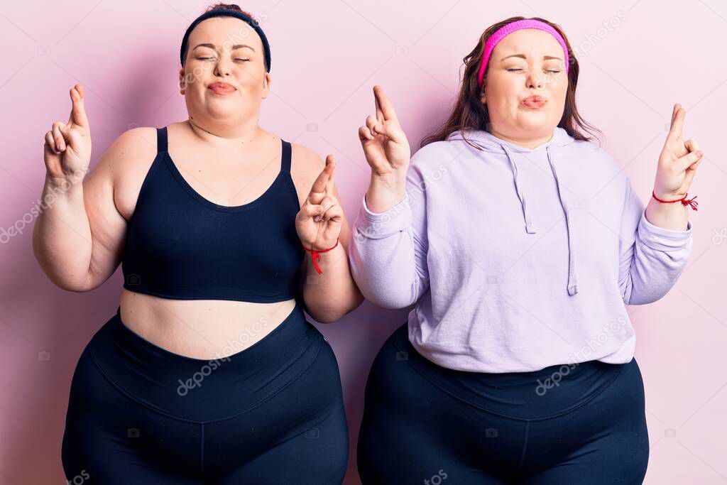 Young plus size twins wearing sportswear gesturing finger crossed smiling with hope and eyes closed. luck and superstitious concept. 