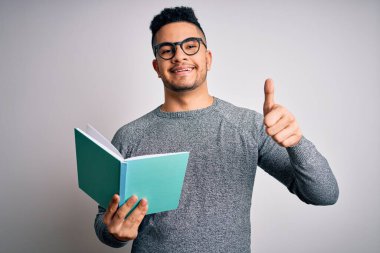 Genç, yakışıklı, zeki bir öğrenci, izole edilmiş beyaz arka plan üzerinde kitap okuyor. Kocaman gülümsüyor ve gülümsüyor. Tamam işareti yapıyor.