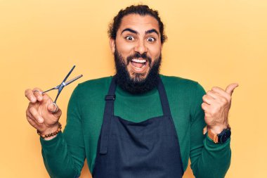 Genç Arap adam berber önlüğü takıyor baş parmağını yana kaldırıyor ve ağzı açık gülümsüyor. 