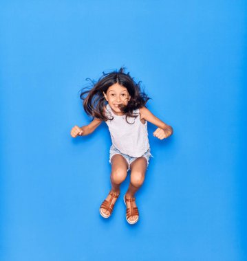 Sevimli İspanyol kız günlük kıyafetler giyip mutlu gülümsüyor. İzole edilmiş mavi arka planda yüzünde gülümsemeyle zıplıyor