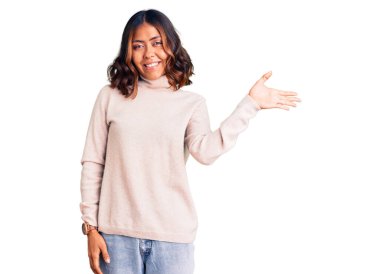 Balıkçı yaka kazak giyen, neşeli bir şekilde gülümseyen ve avuç içi kameraya bakan güzel, melez bir kadın.. 