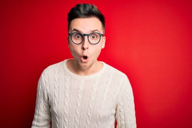 Genç, beyaz gözlüklü ve kırmızı arka planda sıradan bir kazak giyen yakışıklı bir adam korku, korku ve heyecan dolu bir yüz ifadesiyle korkmuş ve şok olmuştu..