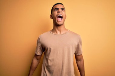 Genç, yakışıklı, Afro-Amerikalı bir adam, günlük tişört giymiş, sarı arka planda dikiliyor kızgın, öfkeli ve sinirli bir şekilde çığlık atıyor, öfkeyle bağırıyor. Öfke ve agresif kavram.