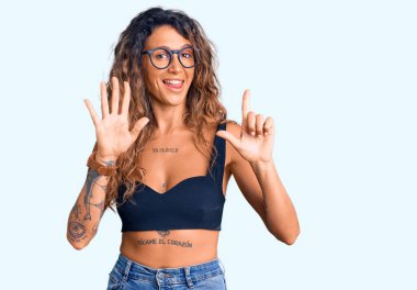 Dövmeli, günlük kıyafetler giyen, gözlüklü genç İspanyol kadın yedi numaralı parmaklarıyla işaret ederken kendinden emin ve mutlu bir şekilde gülümsüyor.. 